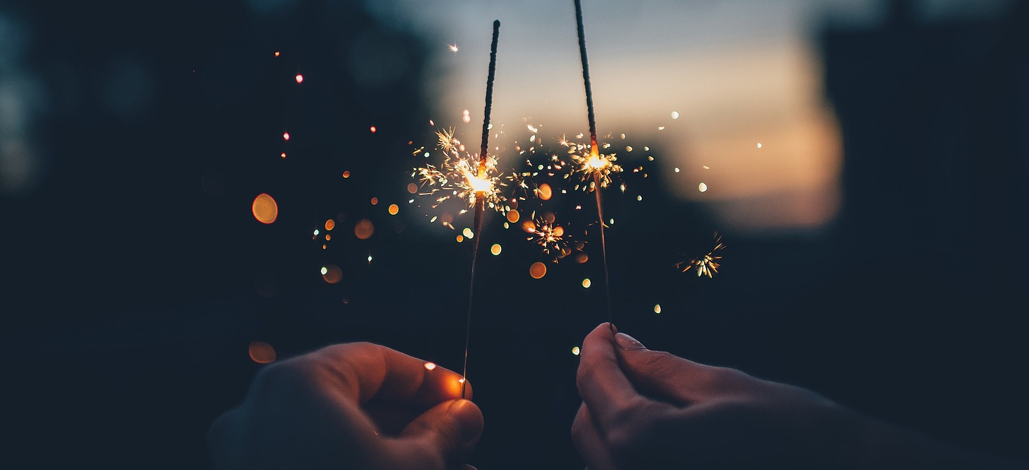 Two hands hold sparklers in the dim evening. The sparks they throw illuminate the hands.