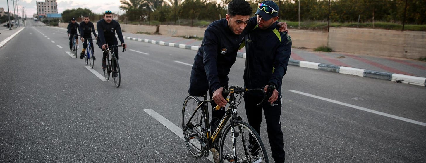A Palestinian para-athlete is supported by another man as he mounts a bike. 3 b i-pedal cyclists trail behind him on the dotted line of an empty road.