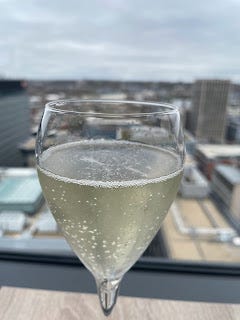 A close up of a sparkling glass of prosecco. It's being held infront of a floor to ceiling window overlooking the skyline of Liverpool. The background view is slightly out of focus as the prosecco is the main thing.