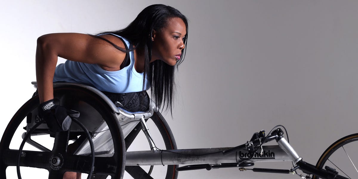 Black woman with long black hair kneels in a wheelchair designed for running competitions. She wears a light blue sleeveless shirt and dark athletic leggins.