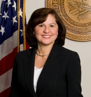 A photograph of a smiling woman in a black blazer with a white shirt directly facing the camera with a U.S. flag and a Justice Department seal behind her
