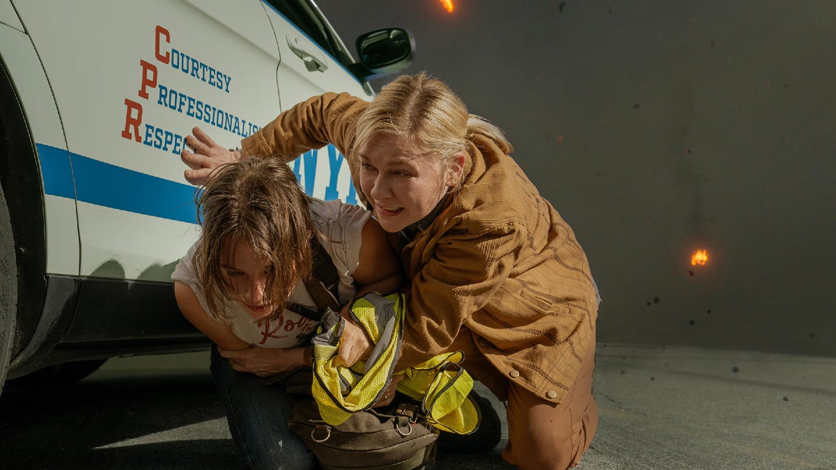 Kirsten Dunst and a civilian kneel beside a car for cover during a shootout scene, highlighting the film's intense action and peril faced by characters.
