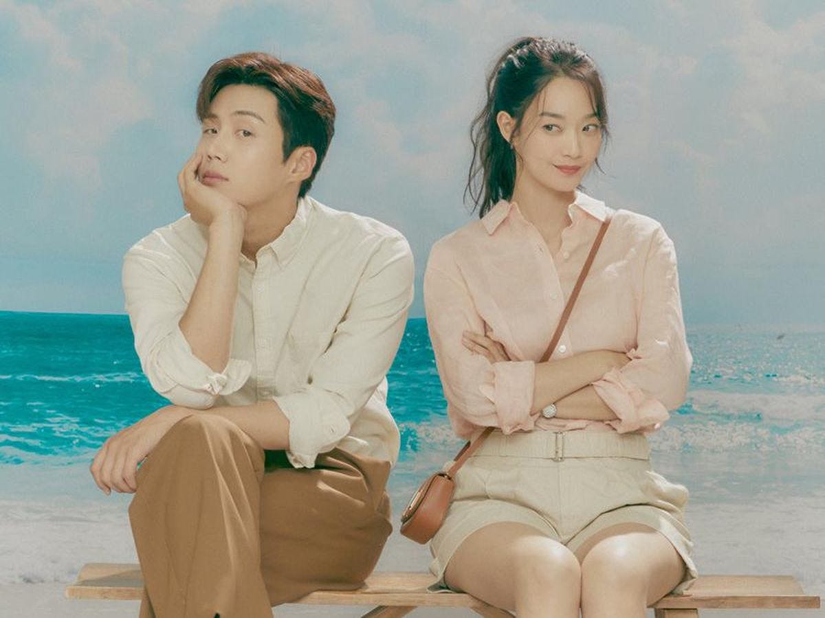 Homem e mulher (Hong Du-sik e Yoon Hye-jin) sentados lado a lado com oceano ao fundo, usando roupas de cor clara em tons terrosos.