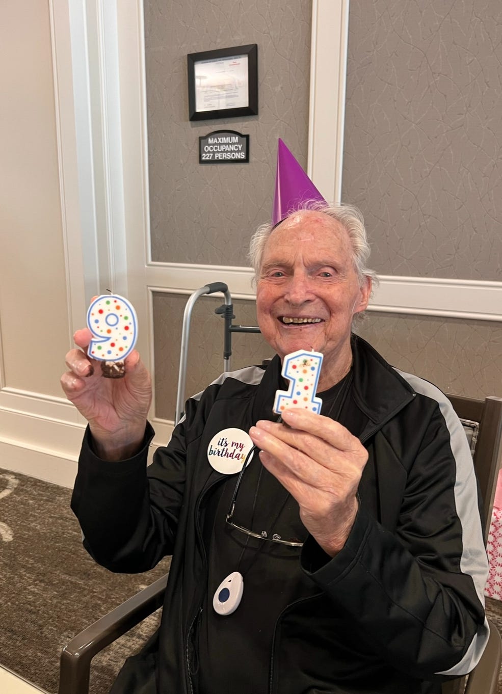 A 91-year-old man holds up two candles, 9 and 1, to celebrate his birthday and smiles at the camera.