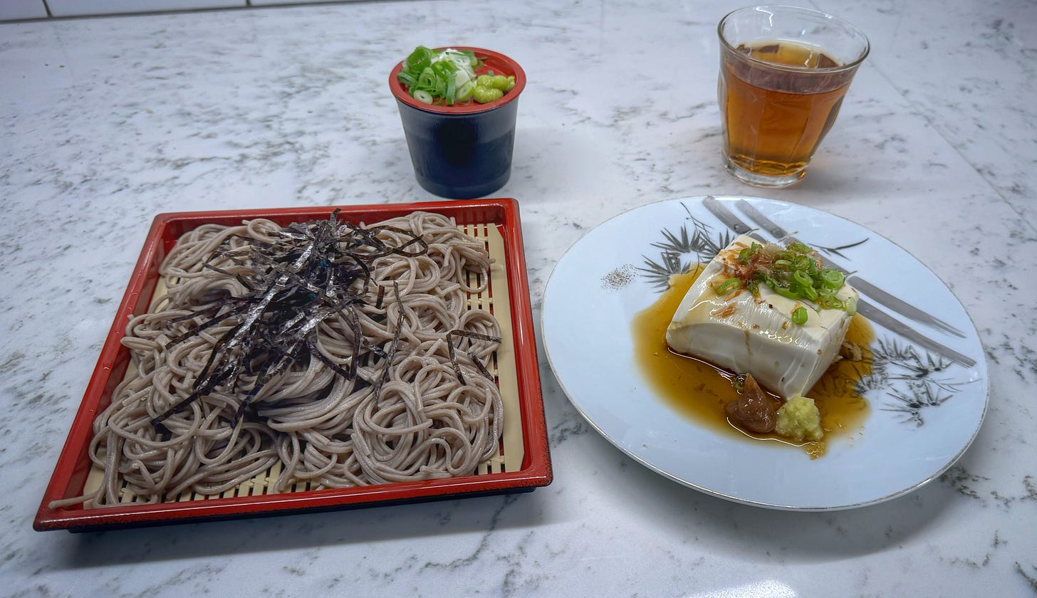 A plate of cold soba noodles with a cup of dipping sauce. On the lid of the dipping sauce there is a pile of green onions and a pile of wasabi. Next to the noodles is a plate with a slab of tofu topped with green onions, bonito shavings, and ponzu sauce. Behind the tofu plate there is a glass of cold, brown tea.