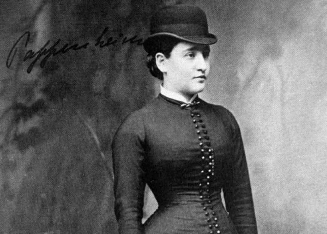 Daguerreotype photo of a white woman with dark hair pulled back wearing a bowler hat and a tight-fitting dress with buttons down the front.