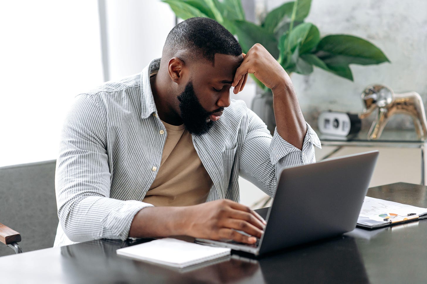 2JPBA7N Sad exhausted african american young man, company employee, sitting at a desk, looks tired and stressed, experience headache, migraine, needs rest, closed his eyes. Overworked from work, deadline