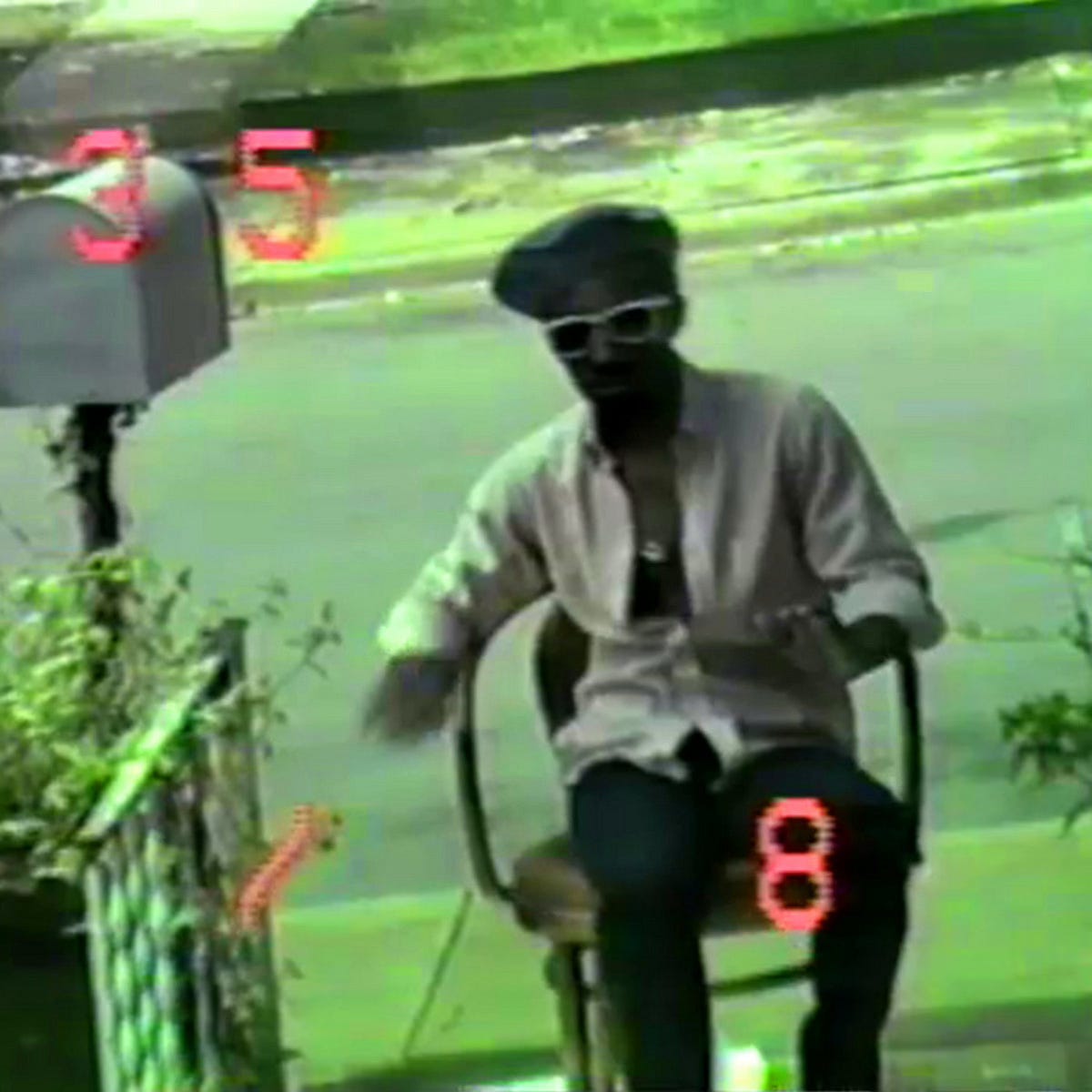 still image from a green-gelled VHS video, featuring a man in a beret and white-framed sunglasses and an unbuttoned white shirt on, arms in motion but seated in a chair near a sidewalk mailbox. The numbers 3, 5, and 8 appear in LCD-style red formatting, perhaps a date stamp from the camcorder.