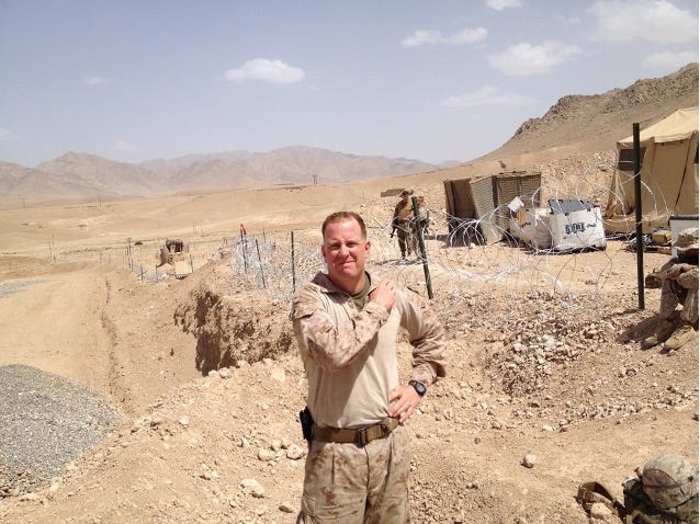 Color photo of the author taken in Northern Helmand, Afghanistan in 2012 during a bright sunny day.  Tim Oliver is in a casual stance in uniform in the center of the photo.  His right arm is across his chest grabbing his left shoulder. In the background to the right of the frame are a tan military tent, and supplies on the ground with a concertina wire fence around the compound. In the distance are mountains against a blue sky with scatter clouds. 