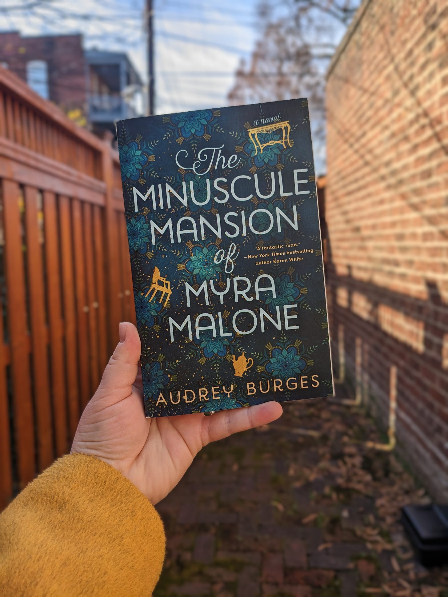 photo of Sarah's hand holding a copy of The Minuscule Mansion of Myra Malone by Audrey Burges in front of a brick wall and fence.