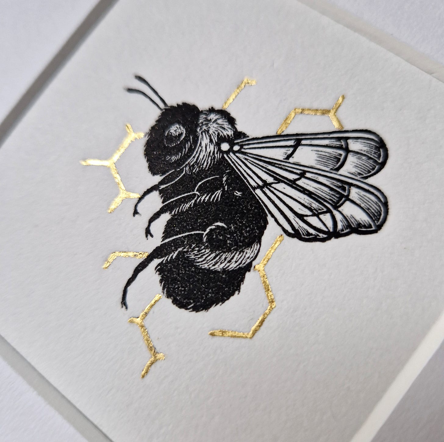 Bumblebee on Golden Honeycomb Wood Engraving. A solitary bee has been printed with black ink on warm white paper against a honeycomb line pattern that has been created using gold leaf.