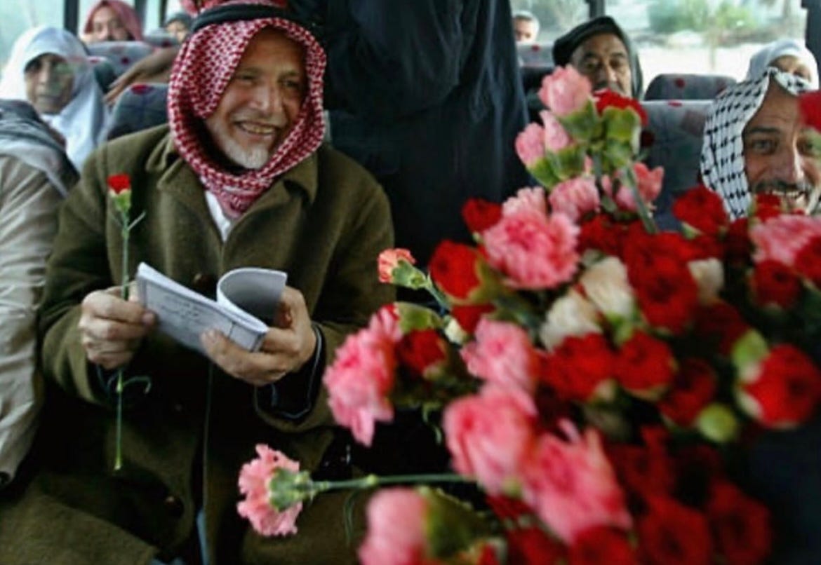 A Palestinian elder smiles, holding an open book and a single flower, riding a bus along with other Palestinians. A bouquet of flowers takes up the foreground 