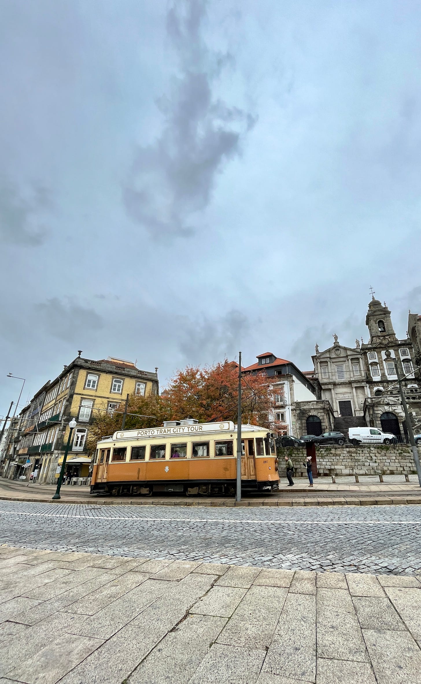 image: a photo of a cheery yellow Porto tram flank by autumn trees, a muted yellow coloured building on the left and the St Francis church on the right.