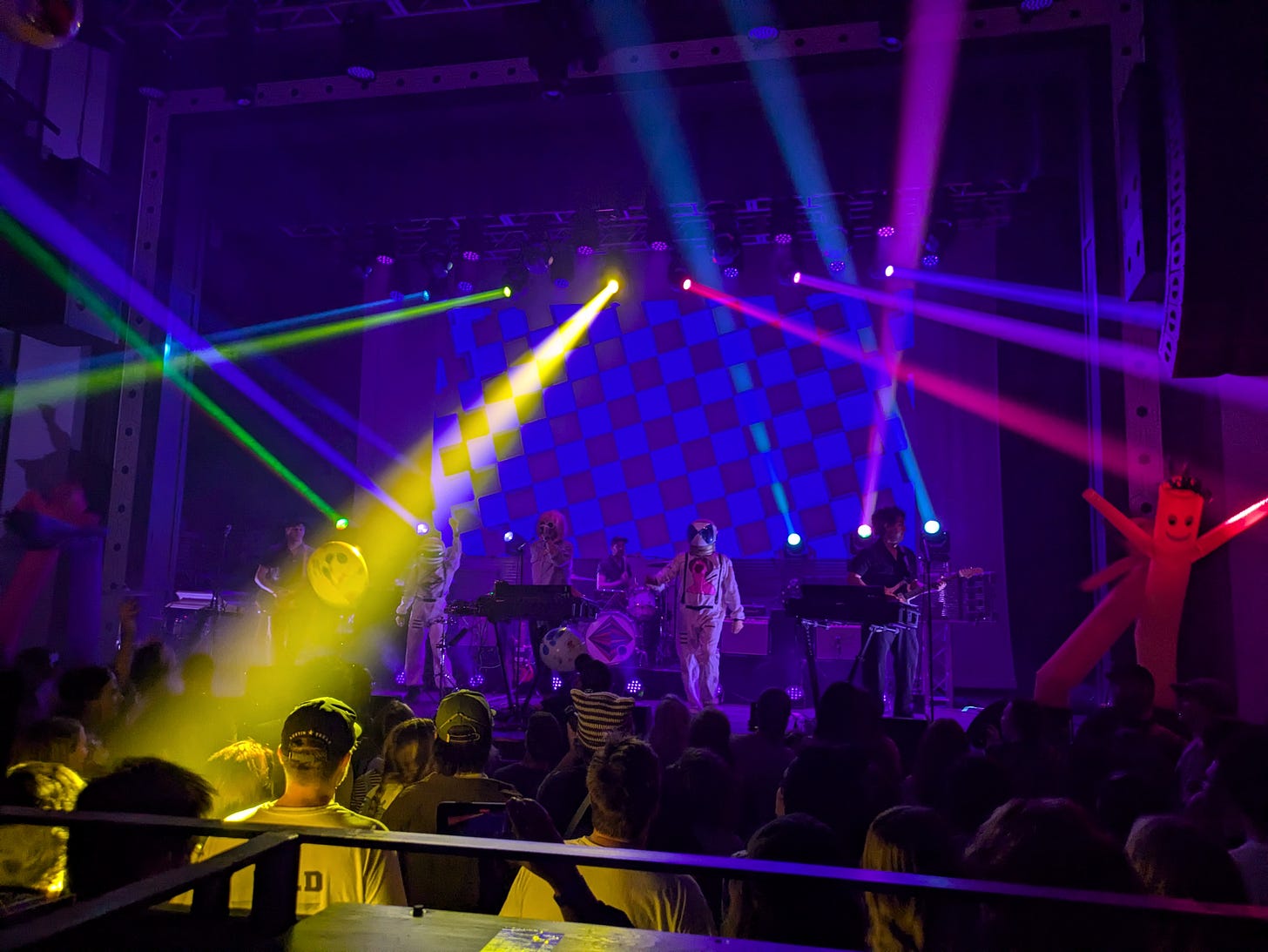 A band on stage. There are two people in astronaut costumes dancing alongside them. Colorful strobe lights flash onto the crowd and wacky inflatable tube men dance on both ends of the stage. A screen behind the band shows a blue and grey checkerboard pattern.