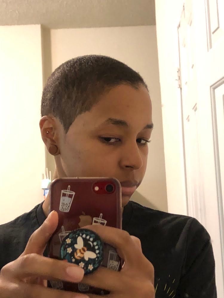 Black girl taking a picture inside a bedroom in front of a mirror looking at new shaved hairstyle with a widows peak prominent