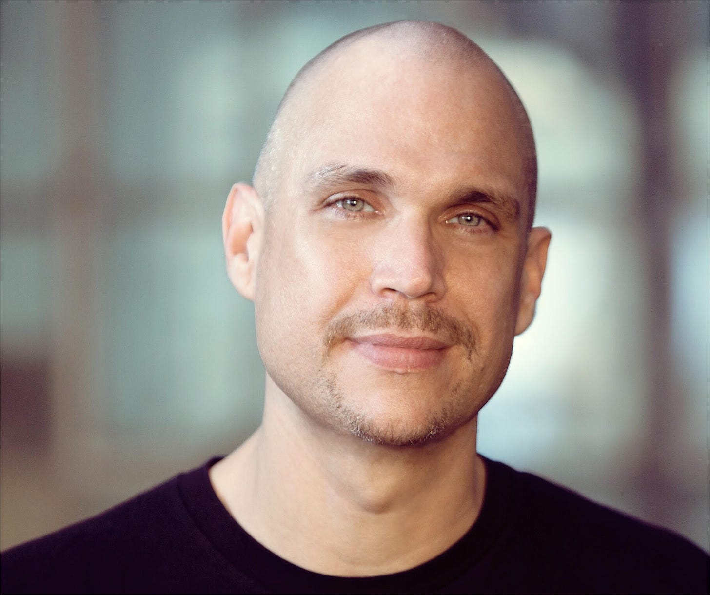 Man with a goatee and light-colored eyes looks a the camera with a slight, closed -lip smile. He wears a black t-shirt.