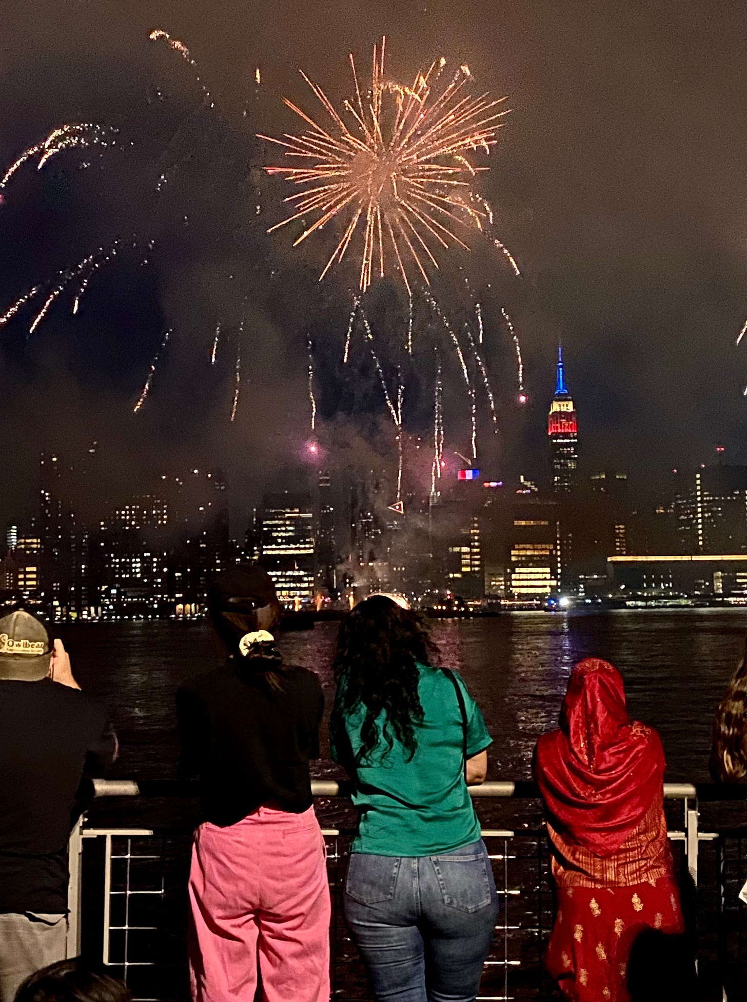 Four New Yorkers leaning on the river rail to watch the 4th of July fireworks. The Empire State Building is visible across the river, light up red, white, and blue.