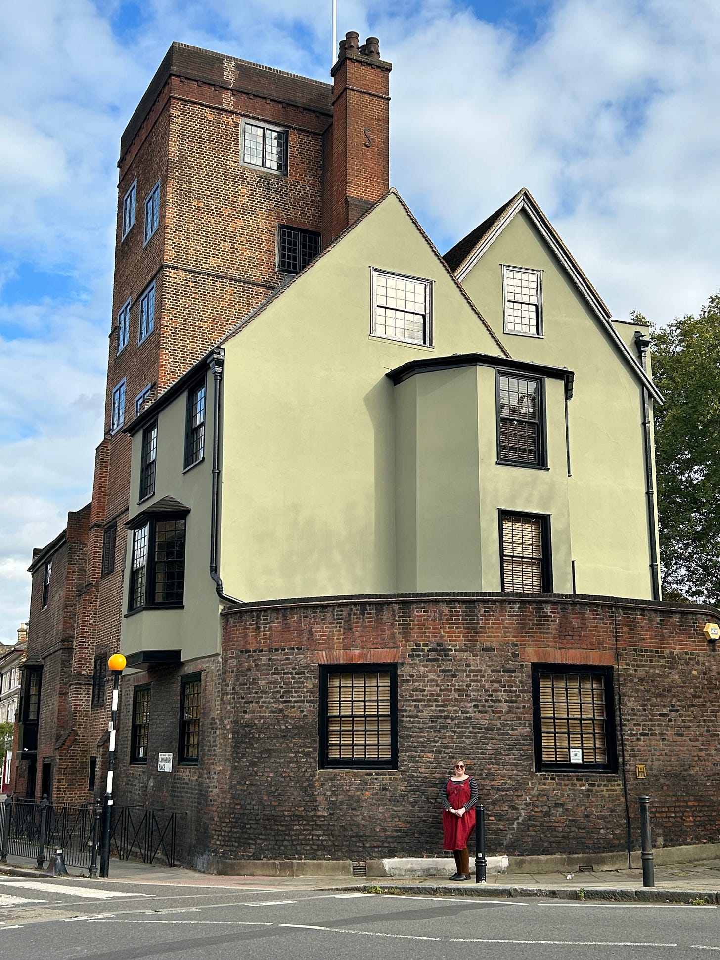 A white woman stands in front of a domestic building with a brick tower. 