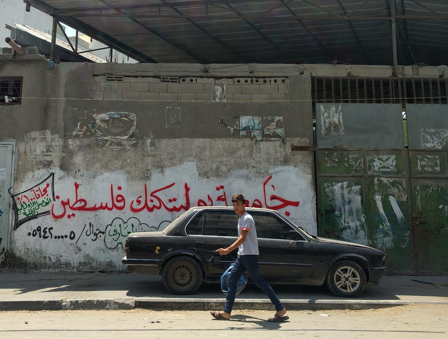 A young man walks past a car in front of a wall covered in Arabic graffiti