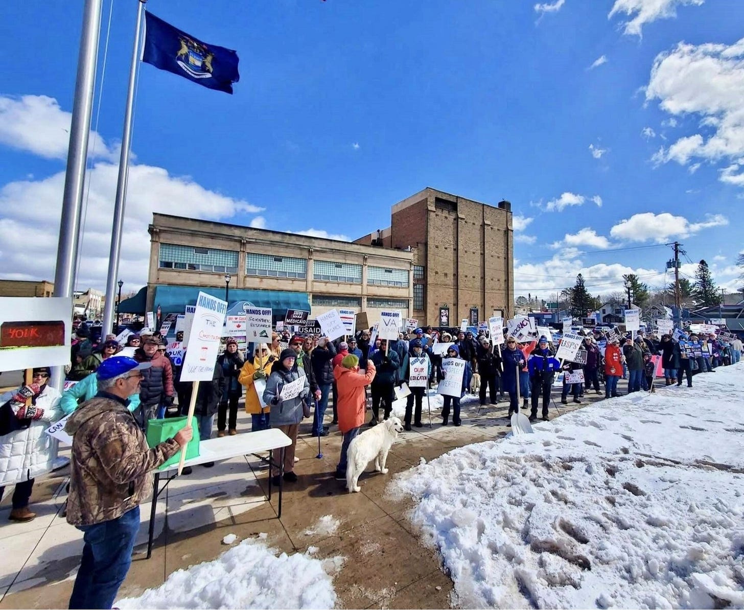 A cold but enthusiastic looking crowd stands on the sidewalk by a Michigan State flag and a commercial building. The sky is blue and there is snow on the ground. Most of the protesters are wearing winter coats and warm hats. They are carrying signs demanding Hands Off USAID, Medicaid, Our Constitution, the VA, and scientific research. A cold but enthusiastic looking crowd stands on the sidewalk by a Michigan State flag and a commercial building. The sky is blue and there is snow on the ground. Most of the protesters are wearing winter coats and warm hats. They are carrying signs demanding Hands Off USAID, Medicaid, Our Constitution, the VA, and scientific research.