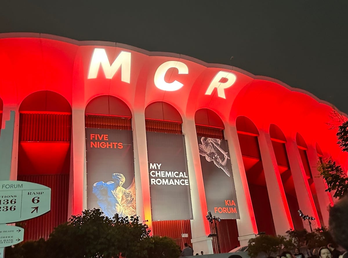 Image ID: The outside of the Kia Forum, a white arena lit up red. 3 black banners with pictured of angels advertise Five Nights of My Chemical Romance. MCR is projected in white against the top of the building. End ID.