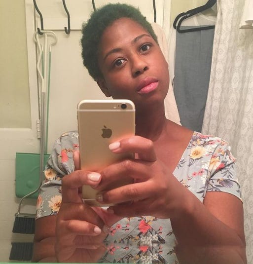 Chanel is taking a mirror selfie with an Apple iPhone. Her hair is green and she is wearing a floral print dress. 