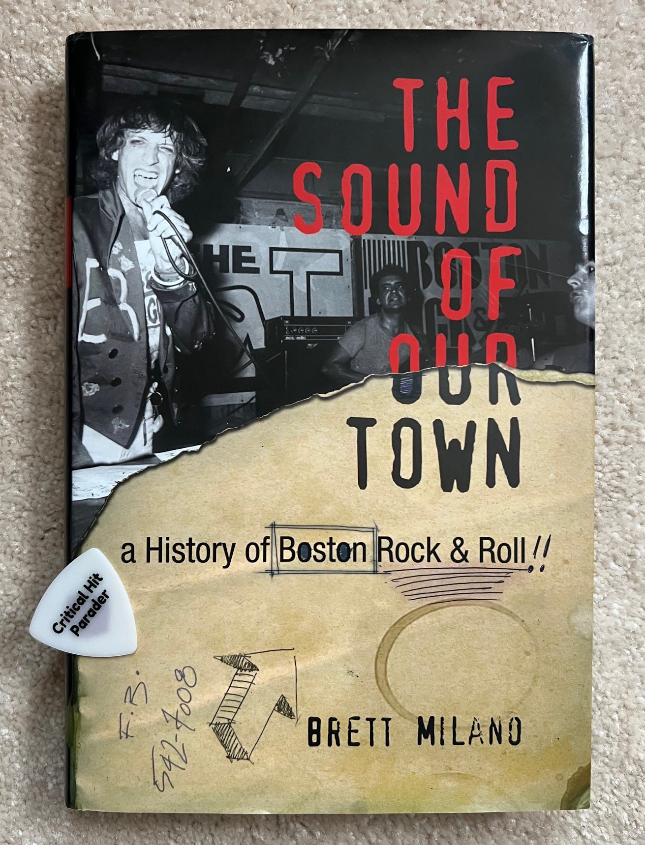 Hardcover book called The Sound of Our Town - a History of Boston Rock & Roll by Brett Milano. It features a photo of Willie Alexander singing on stage. A white Critical Hit Parader guitar pick sits slyly on top of the book.