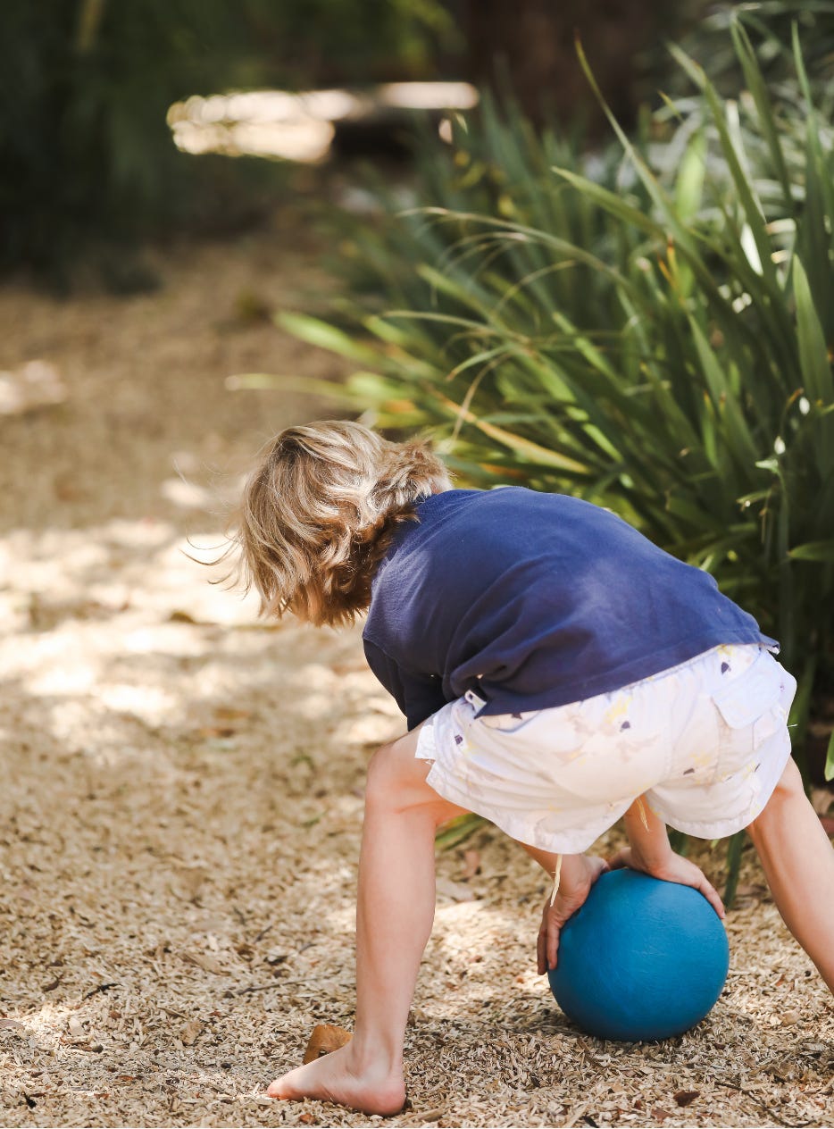 Looking at the back of a small child who is bending over to pick up a blue ball. The child is wearing white shorts and a blue T-shirt and has bare feet. They're on a sawdust path and there are some plants on the right and top of the image.