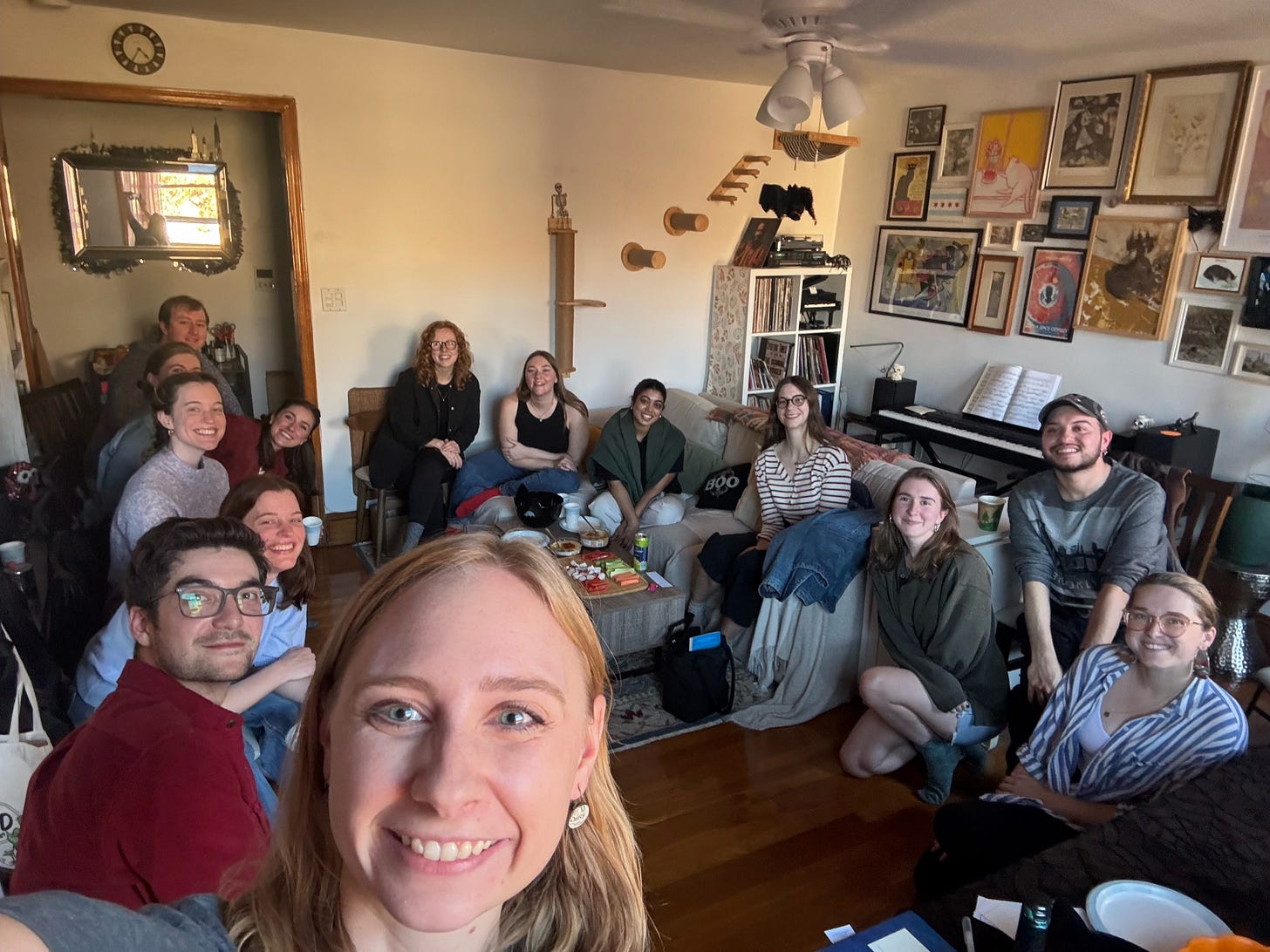 A group of fourteen people sitting in a living room on chairs and couches in a circle, smiling.