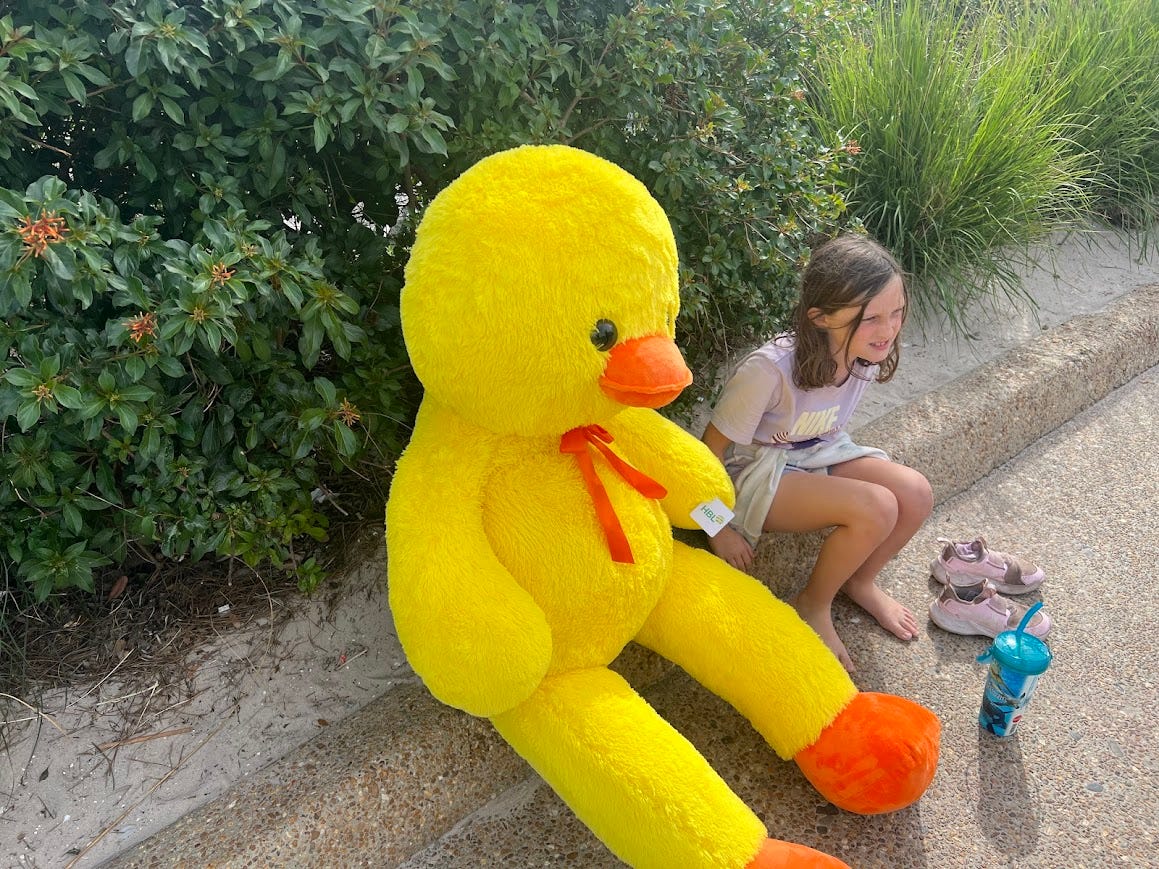 Photo the giant duck sitting on the curb of a sidewalk next to the author's daughter.