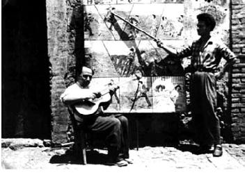 foto in bianco e nero di un cantastorie seduto con una chitarra con un fondale dipinto che raffigura le vicende dei paladini di francia