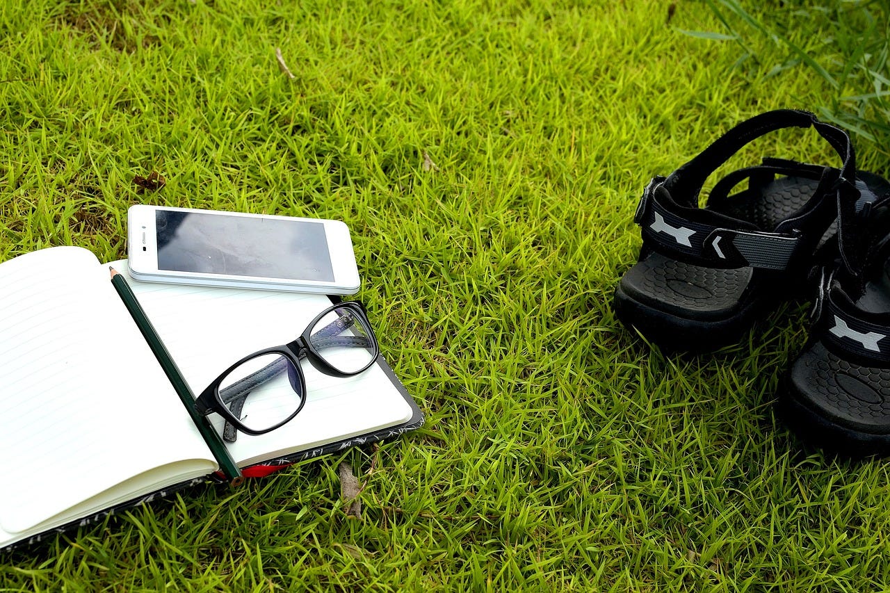 A patch of grass on which a journal, a pencil, eyeglasses, a phone, and a pair of sandals rest.