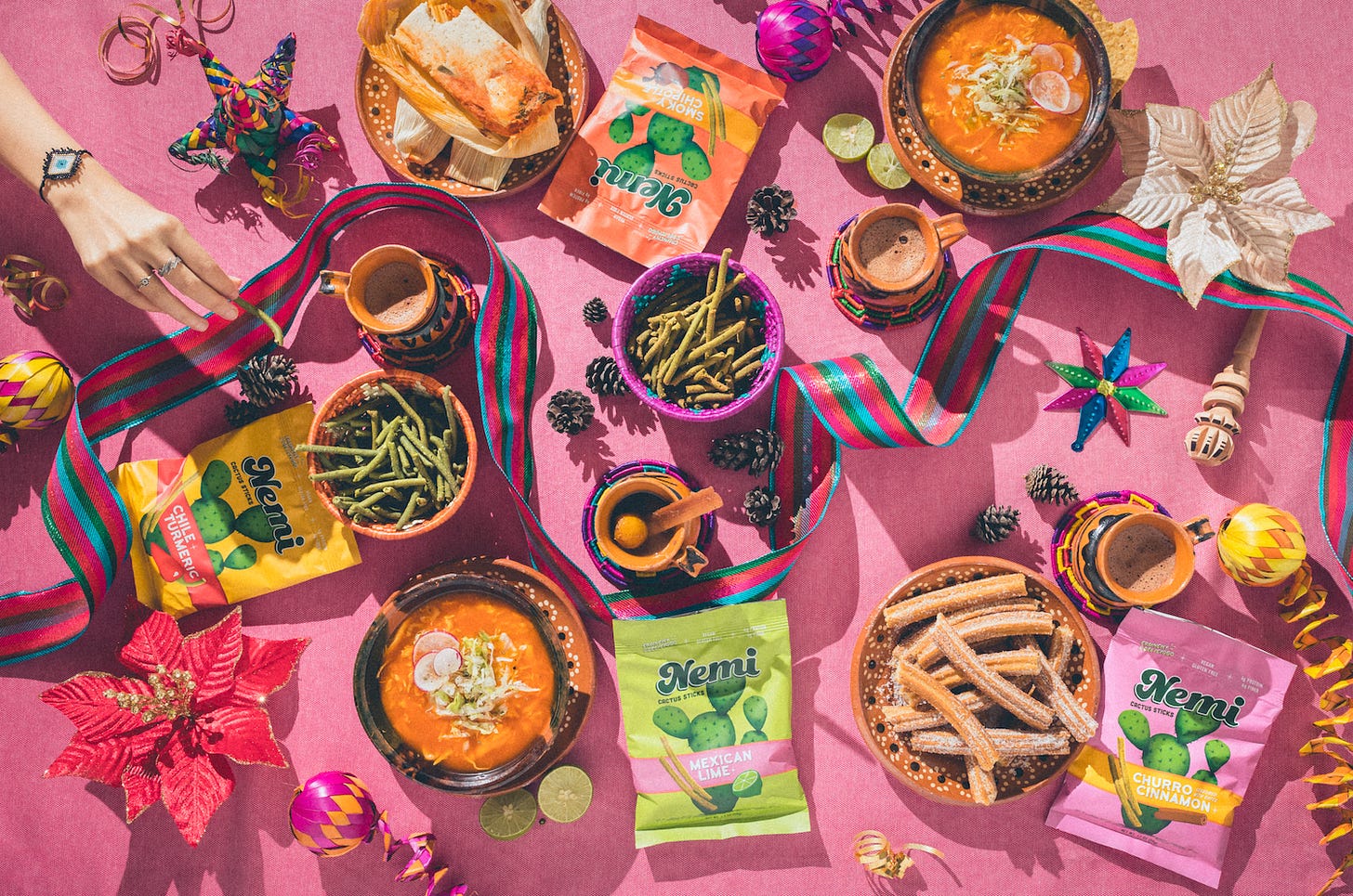 Colorful table with bags of Nemi Snacks nopales snack bags in chile-turmeric, Mexican-lime, smoky-chipotle, and, churro-cinnamon flavors surrounded by Mexican food and dips on a pink tablecloth surrounded by brightly colored traditions Mexican decorations.