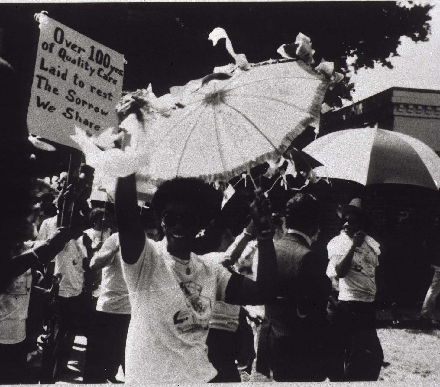 The black community of New Orleans in a black and white photo dated from the 1980's staging a jazz funeral and protest about the closing of a public hospital. Participants of the protest are in the background with umbrellas raised. One is carrying a sign that reads: "Over 100 years of Quality Care Laid to rest The Sorrow We Share".