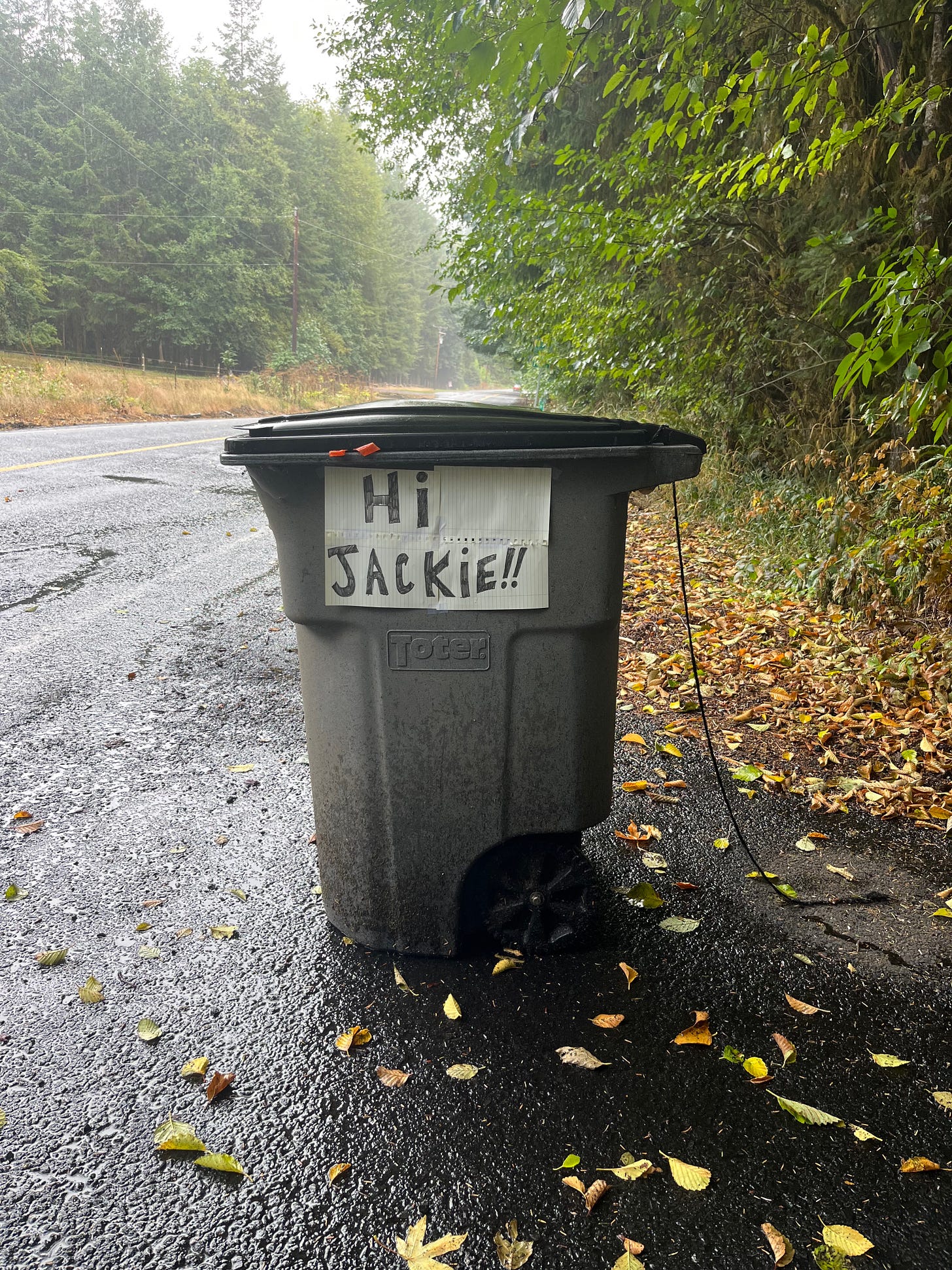 A black garbage bin on the side of the road with a sign taped to it that reads "Hi Jackie!!"