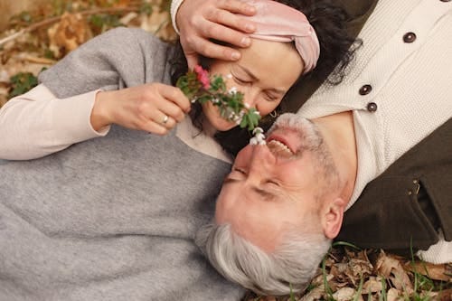 Free From above of cheerful senior wife wearing wide scarf and headband with flower bouquet in hand and happy elderly gray haired husband in warm clothes lying on ground with fallen leaves in park with closed eyes Stock Photo