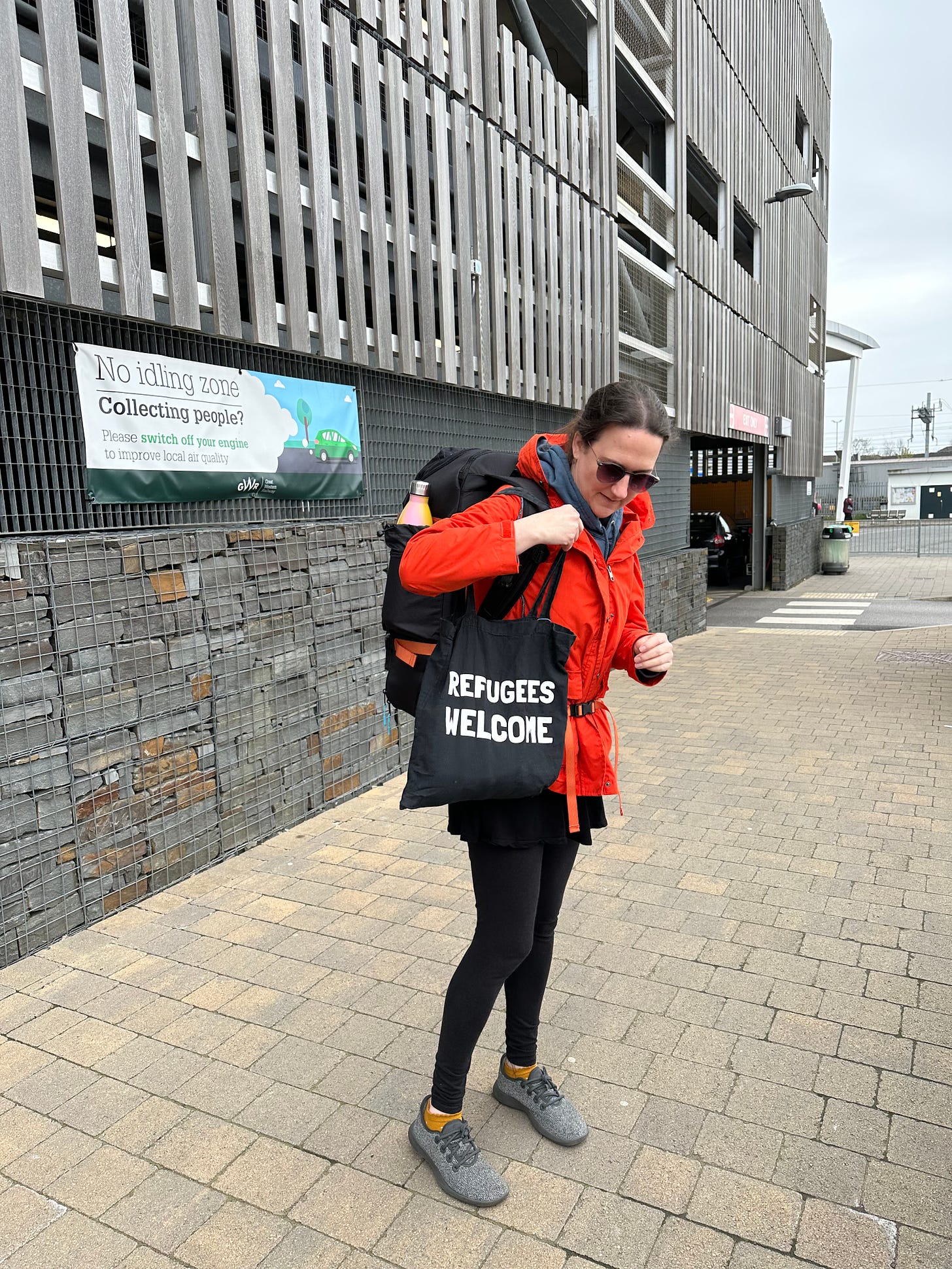 I am standing outside what is a 70s ? train station wearing sunglasses and a bright orange coat and black leggings and a black backpack and I am holding a black tote bag that says refugees welcome on it. 