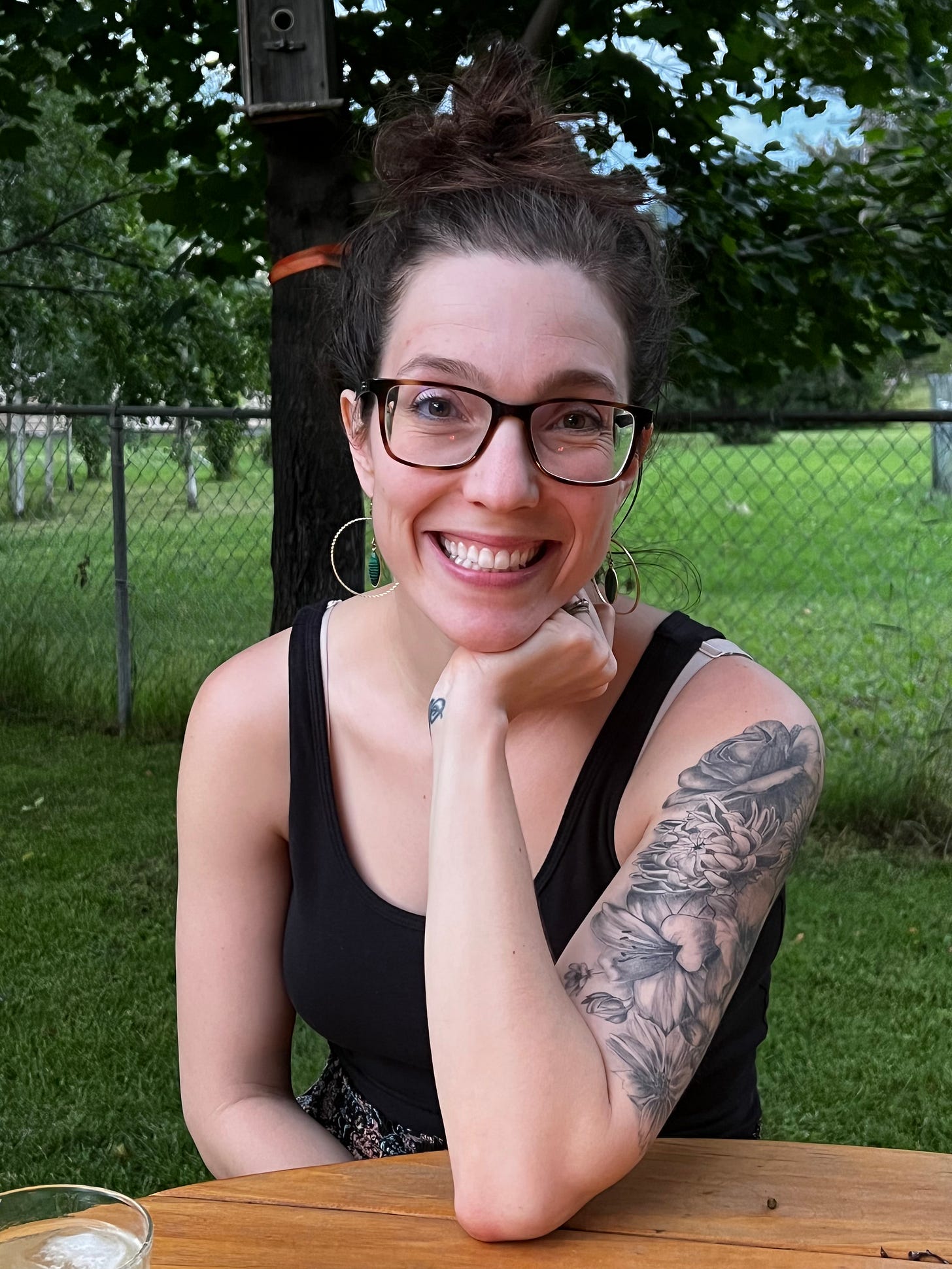 A smiling woman with glasses and a floral tattoo on her arm sits at a wooden table outdoors. She rests her chin on her hand, with greenery in the background.
