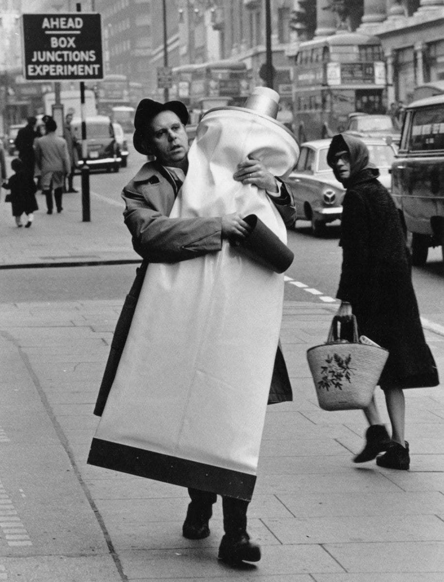 A busy street in London. A man, facing us in a trenchcoat and a dark hat, mouth open as if breathing heavily, has his arms wrapped around a human-sized toothpaste tube. He carries the cap in his right hand. A woman in a kerchief turns to gawp at him!