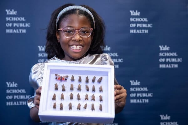 Bobbi Wilson smiles as she holds up a display with 27 spotted lanternflies, preserved from ones she caught in her neighborhood in Caldwell, New Jersey. One of the lanternflies has its wings spread out, showing orange, black and brown colors.