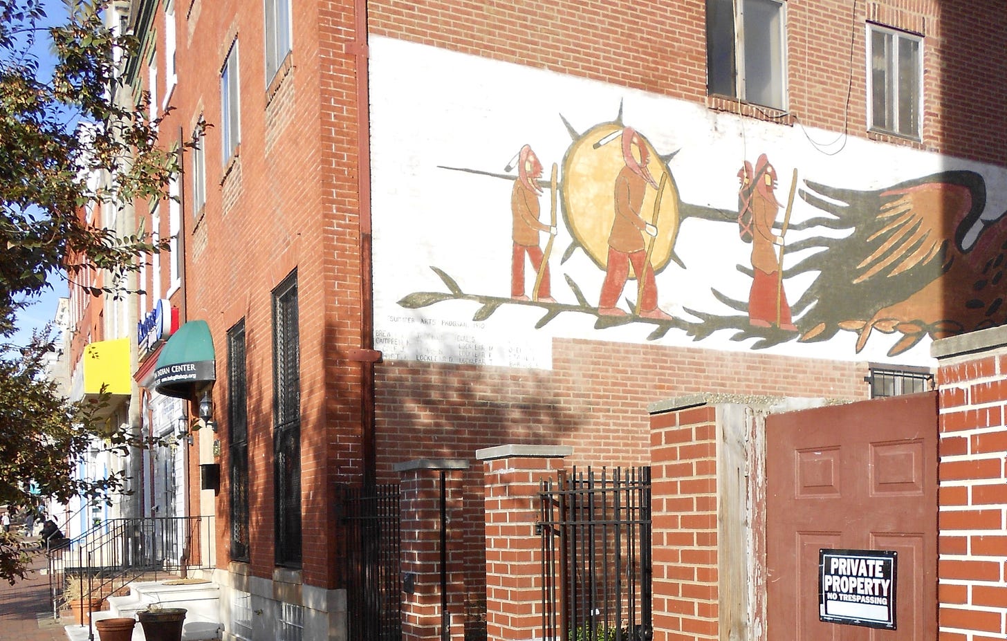 Photograph taken from the street of the side of the Baltimore American Indian Center. It features a painted mural on the side of the building, containing traditional imagery.