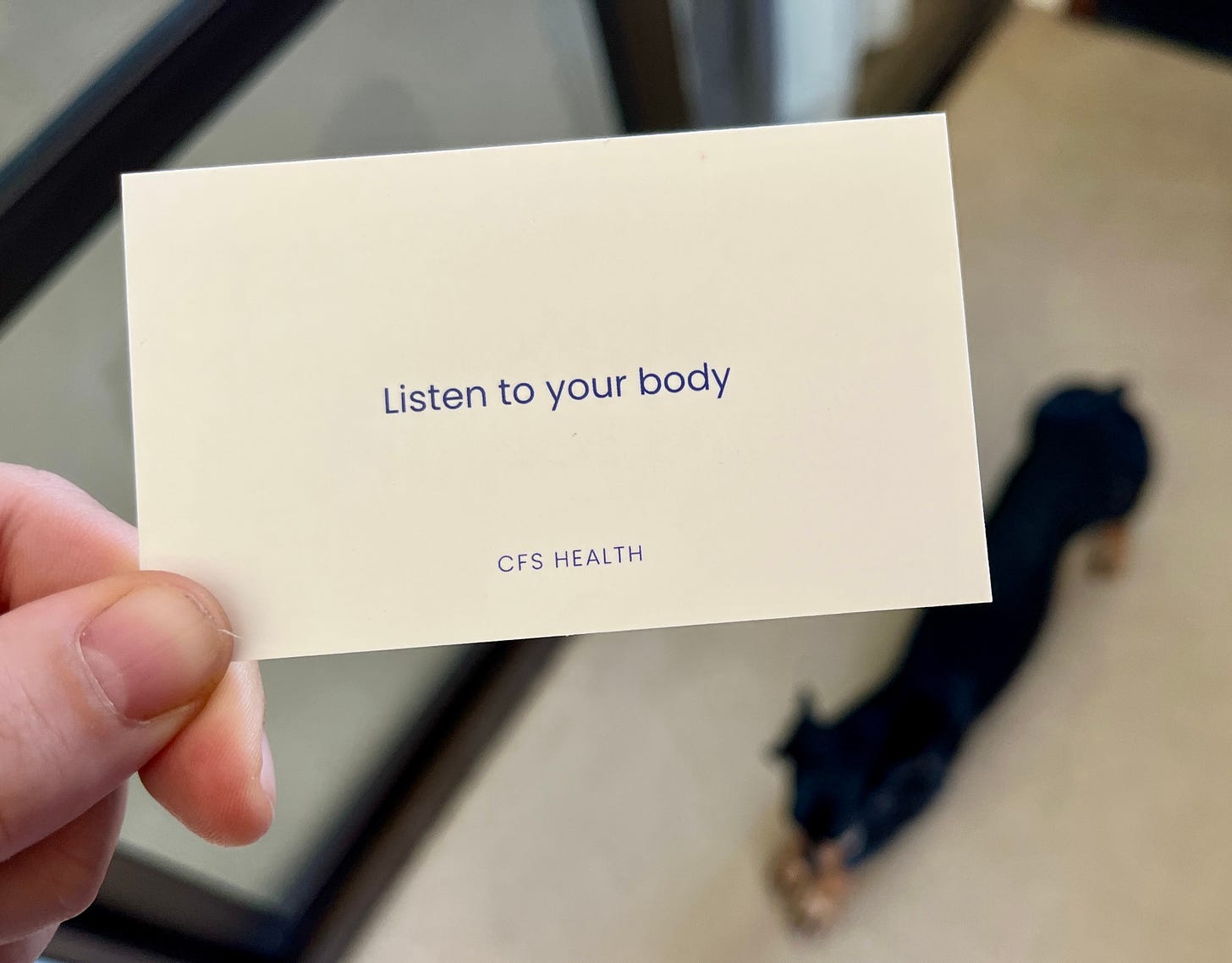 in the foreground a hand holds a card that reads  "listen to your body" with a logo that reads "CFS health". In the blurry background a black dachshund does a stretch.