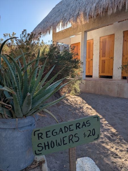 a hand-printed sign advertising showers in Spanish and English for 20 pesos--one dollar--with a white building with a thatched roof and wooden doors on individual shower stalls