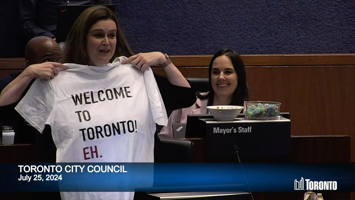 Councillor McKelvie holds a white t-shirt reading "Welcome to Toronto eh" in the council chamber