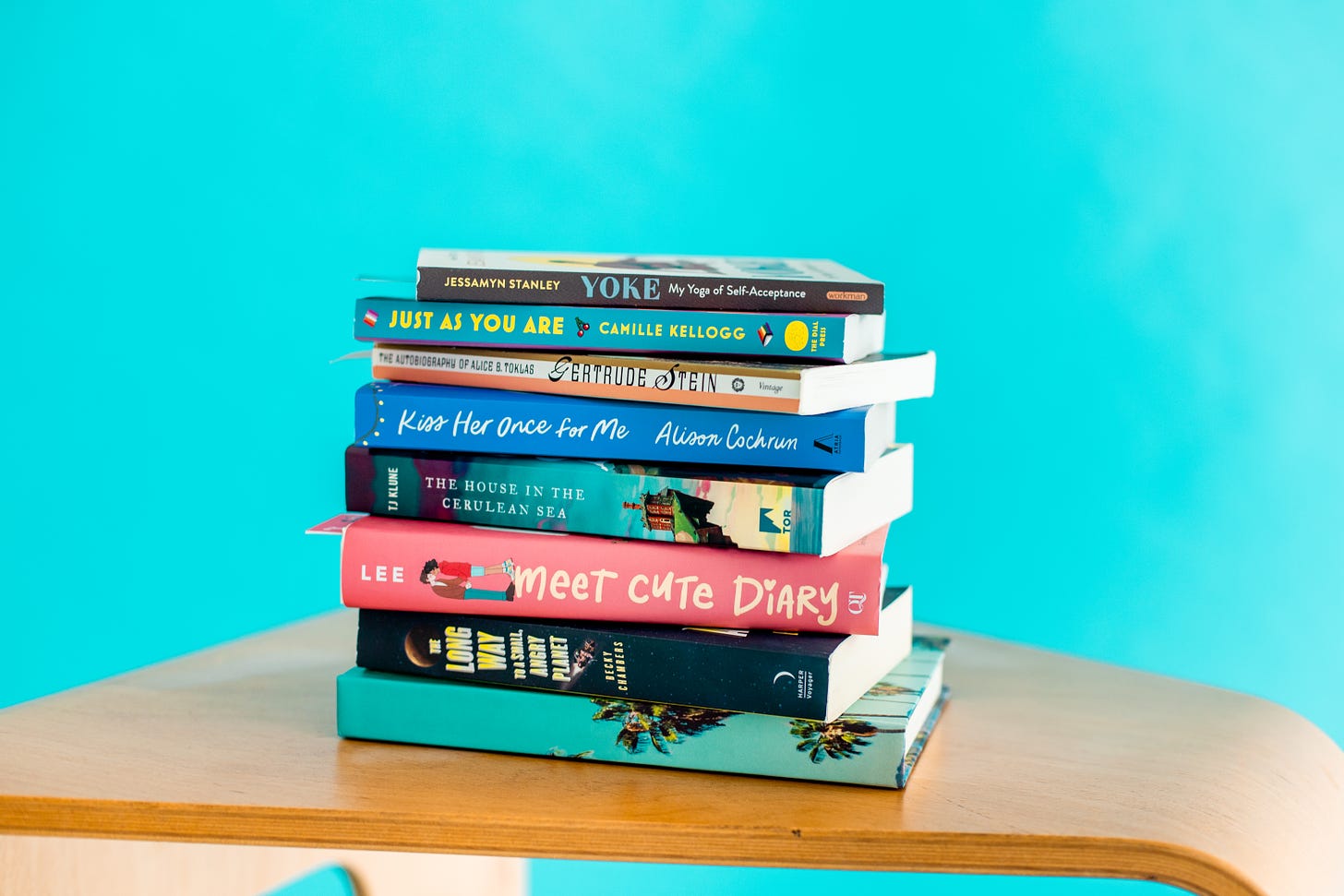 A stack of books by LGBTQ authors including Becky Chambers, Emery Lee, TJ Klune, Alison Cochrun, Gertrude Stien, Camille Kellogg, and Jessamyn Stanley.