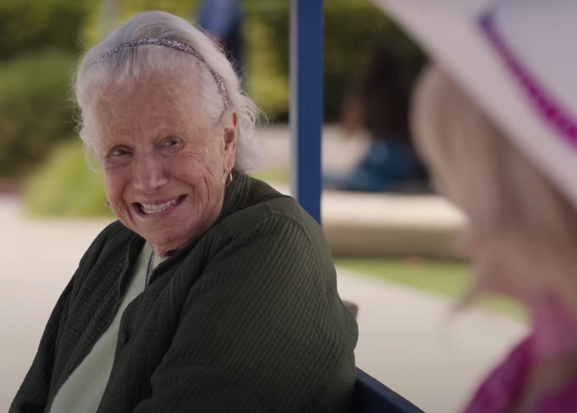 Scene from Barbie movie with older woman smiling on bench.