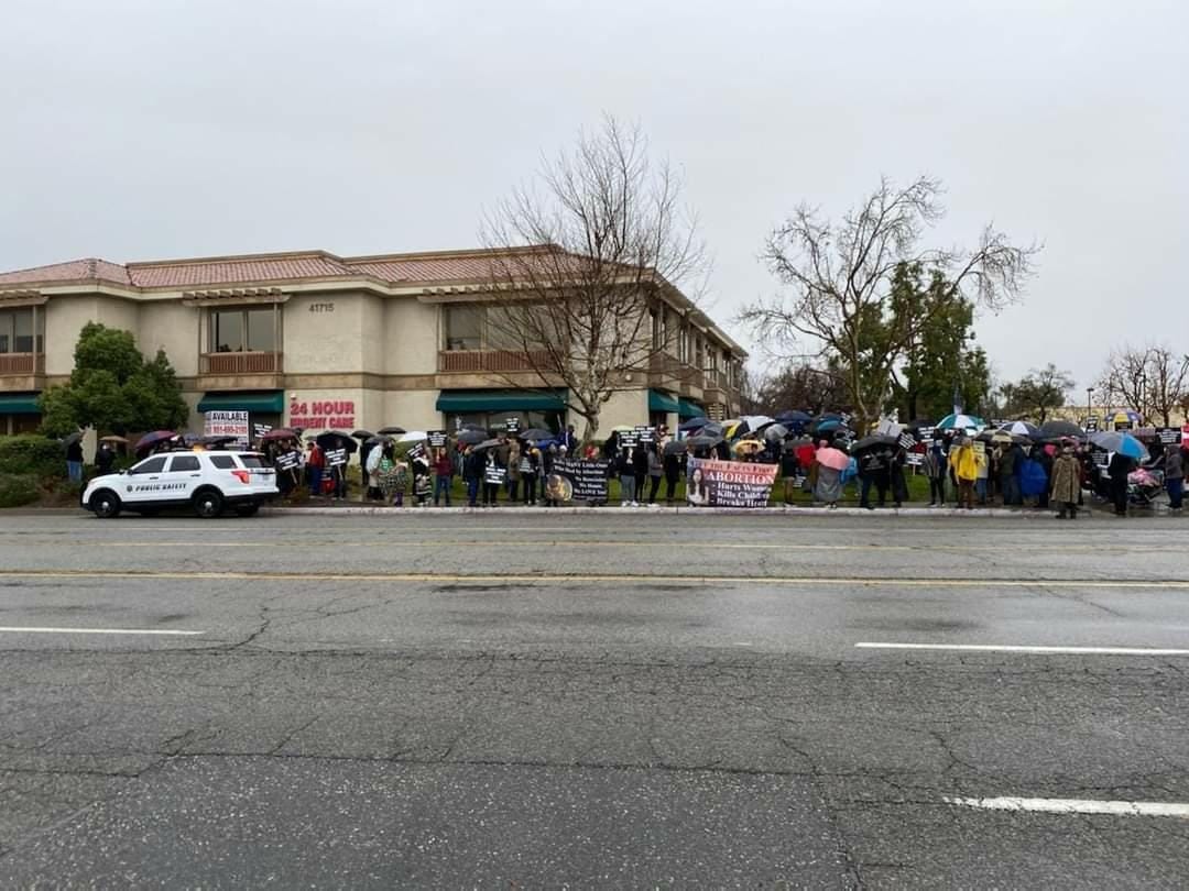 Participants of a March for Life outside of an abortion clinic standing and walking in the cold rain under a gray sky.