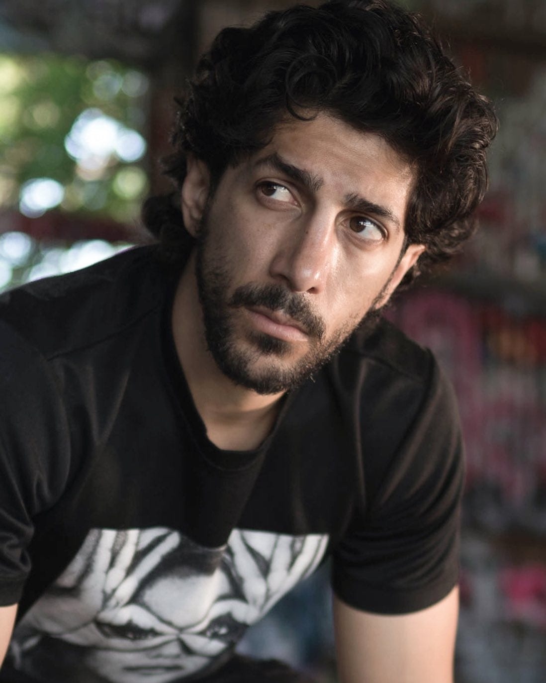 A portrait of Michael Angelo Zervos, a man with dark curly hair and facial hair, wearing a black tee with white print