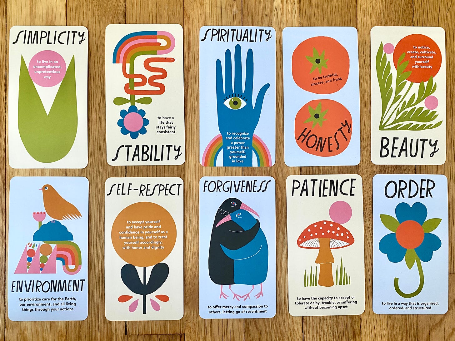 Ten colorful values cards sitting on a wood floor. Each card names a value, which includes simplicity, stability, spirituality, honesty, beauty, environment, self-respect, forgiveness, patience, order. Each card has the name of the value and a colorful illustration by Lisa Congdon that represents the value.