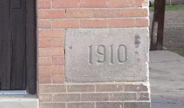 A large grey stone with the year 1910 pressed into it is shown set into the side of a brick building.